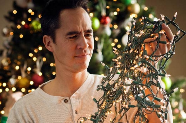 Man stood in front of a Christmas tree holding tangled Christmas lights  and looking confused. 
