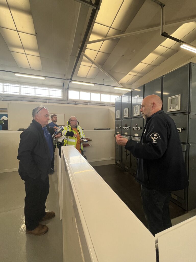 Phil stands in front of PASS' mock substations explaining testing processes to the attendees gathered around the entrance to the mock substations. 