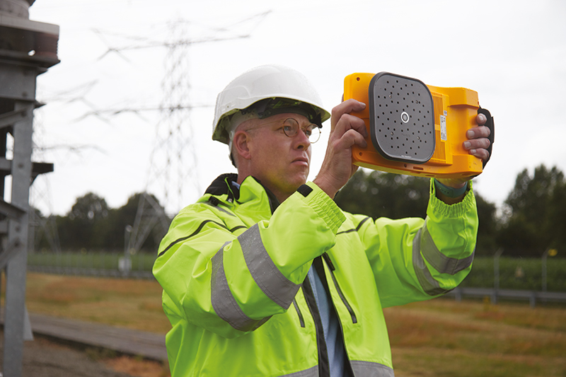 Man in high-vis and white hard hat using the ii910 outside to detect faults. He holds the ii910 in front of him; the back of the unit with the microphone array is visible. 