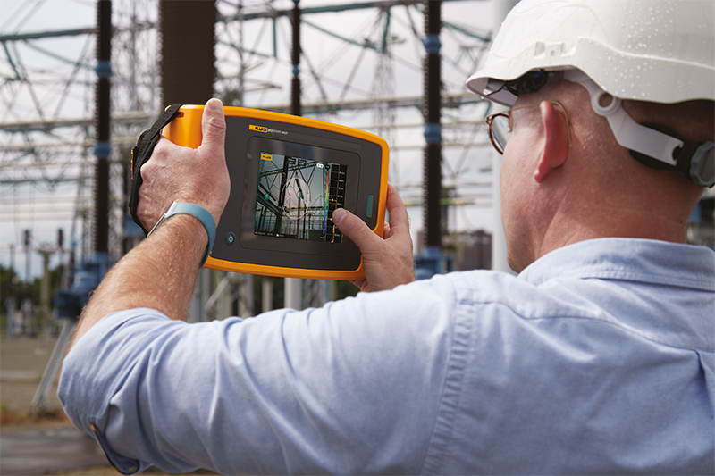A man in a white hard hat is using the Fluke ii910 to inspect high-voltage electrical equipment. He stands at a safe distance with the Fluke ii910 in front of him. The touchscreen display is visible. 