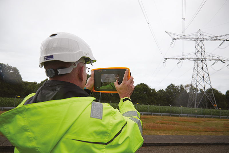 A man in high-vis and a white hard hat is using the Fluke ii910 to check a pylon. He stands at a safe distance with the Fluke ii910 in front of him. The touchscreen display is visible. 