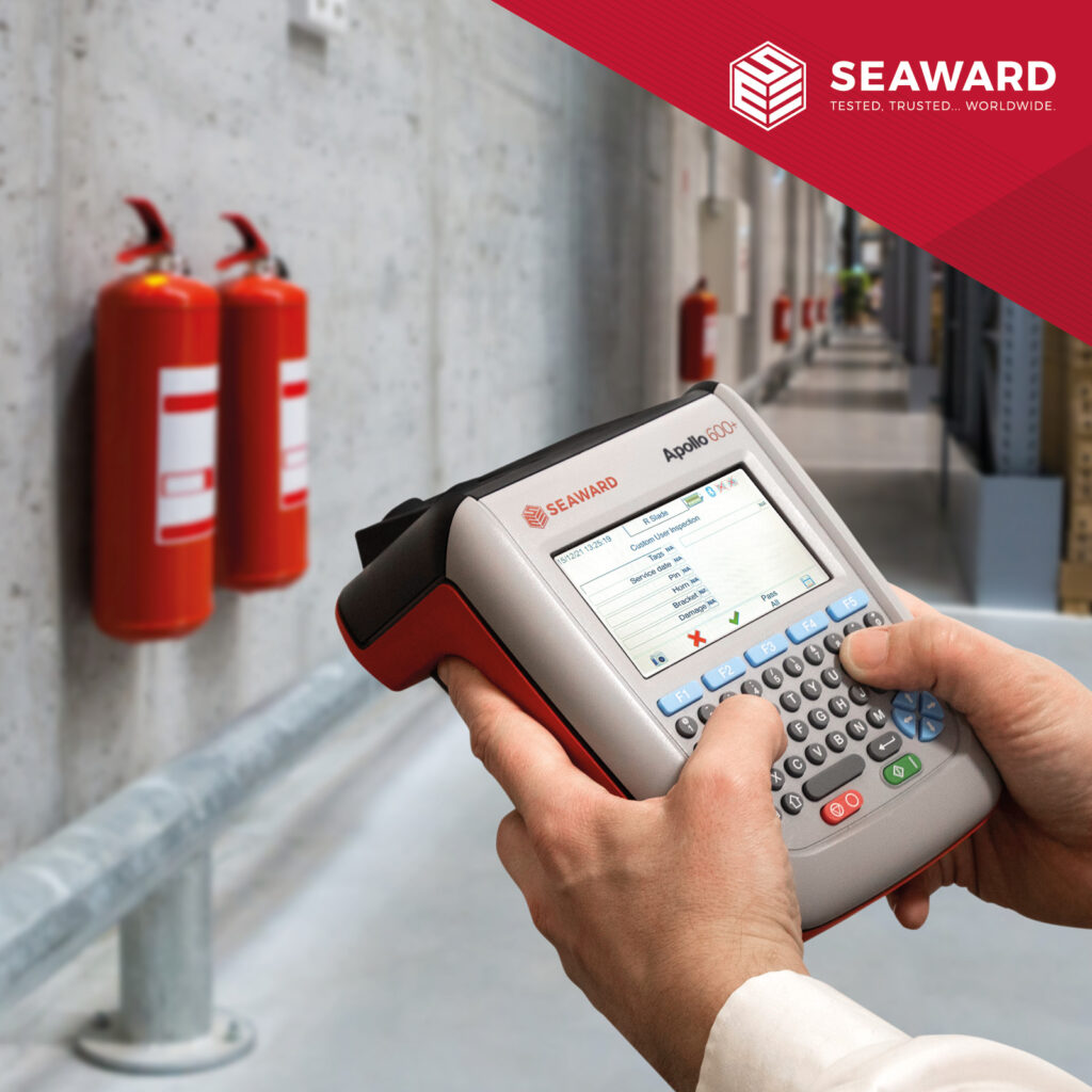Some hands hold a Seaward Apollo+ PAT Tester.  A corridor with red fire extinguishers is visible in the background. the Seaward logo sits on a red background in the top right of the image. 