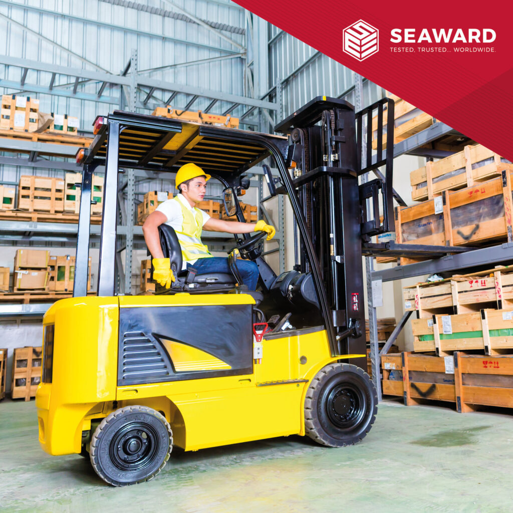 A man in yellow high-vis and a hardhat sits on a yellow forklift truck in warehouse with shelves full of wooden boxes. The Seaward logo sits on a red background in the top right corner of the image. 