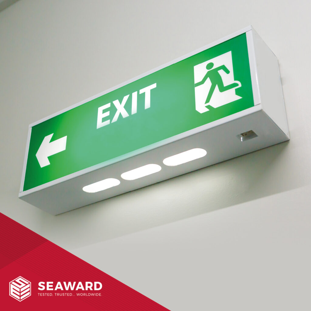 An illuminated, green emergency exit sign on a white wall. The Seaward logo is in the bottom left of the image. The logo is on a red background. 