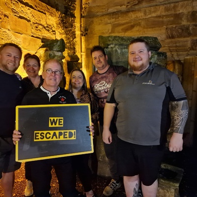 Six members of the calibration department pose for a photo in the castle dungeons. Paul stands in the middle holding a sign saying 'We Escaped!'