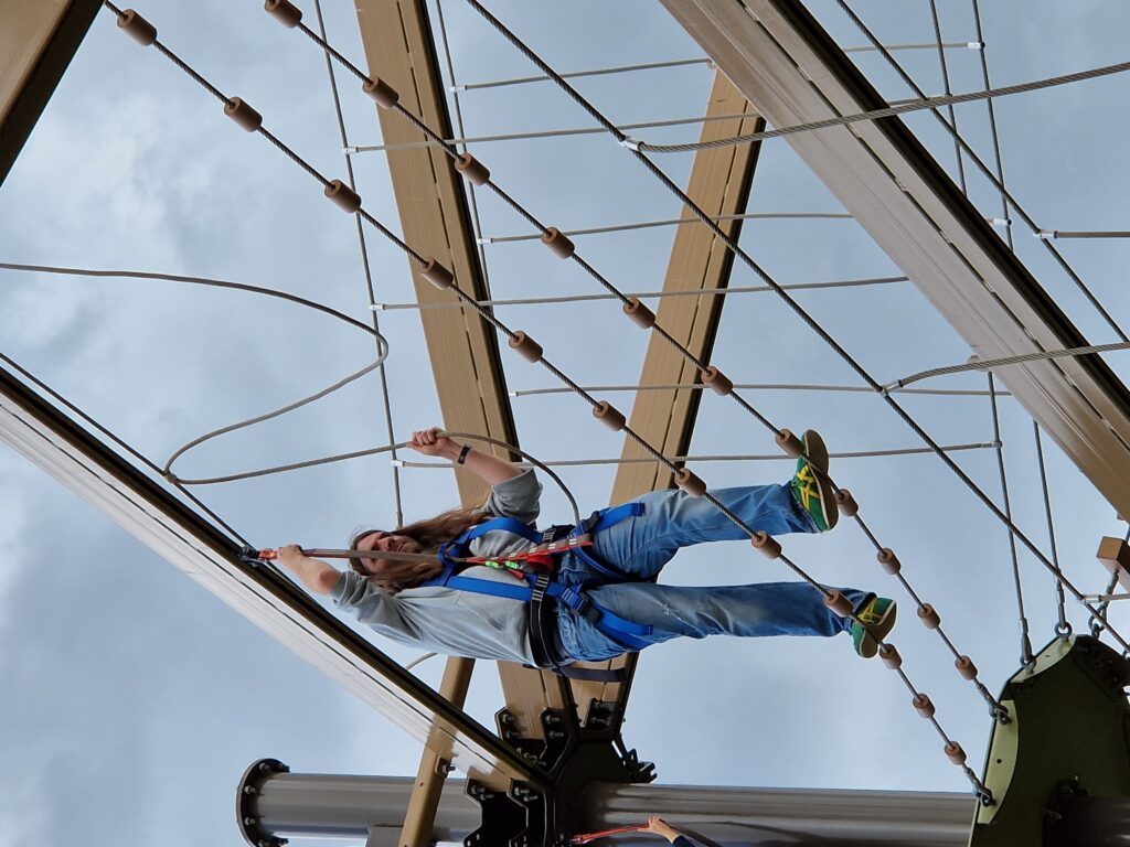 Stacey crosses a bridge consisting of two parallel ropes with evenly spaced cylindrical wooden markers. He holds onto his harness and a loose, looping rope handrail. 