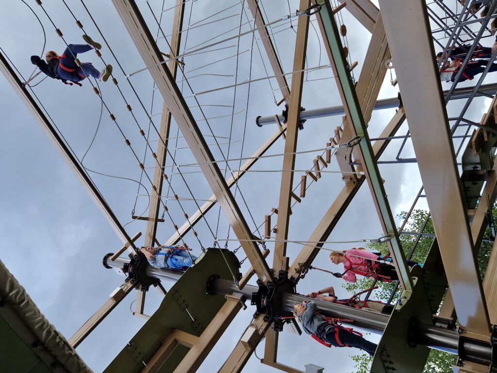A photo taken from beneath the Air Trail. Rachael crosses a bridge consisting of two parallel ropes with evenly spaced cylindrical wooden markers.  Stacey watches on from the pillar and platform behind her. On the level immediately below, Jess, Katie and Matt look up at Rachael. 