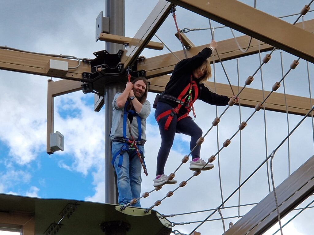 Stacey stands against a pillar clinging to his harness, while a smiling Rachael crosses a bridge consisting of two parallel ropes with evenly spaced cylindrical wooden markers.  