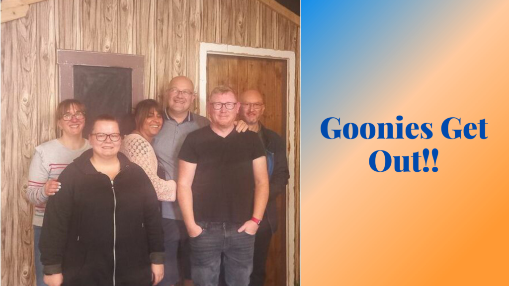 On the left of the banner is an image showing three men and three women smiling in front of a wooden door and wall. To the right of this large, blue text on an orange and blue background reads "Goonies Get Out!!"