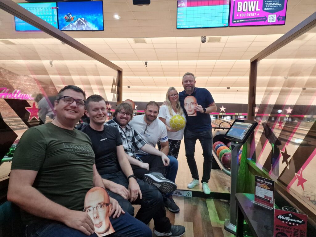 The sales team gather together for a photo next to their bowling alley. Chris M, Chris C, Lewis and Gary (holding a cut-out of Steve's head) sit while Claire holding a yellow bowling ball and Dave holding a cut-out of Steve's head stand at the back. 
