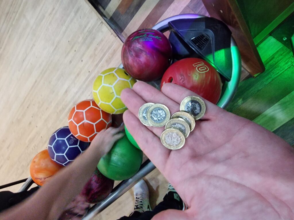 A hand holding £1 is held out over a collection of bowling balls in a variety of colours. 