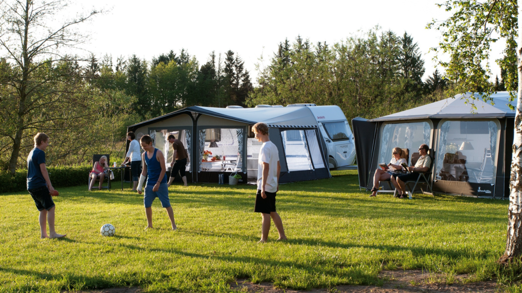 Some teenagers play football on a field. Behind them, parents sit on deck chairs in front of caravans and large tents with clear walls. 