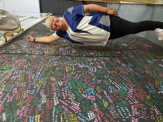 Dawn, dressed in a blue and white top and black trousers, signs her name on the 'Wall of Clutch'. A long blackboard with names written in different coloured chalks. 