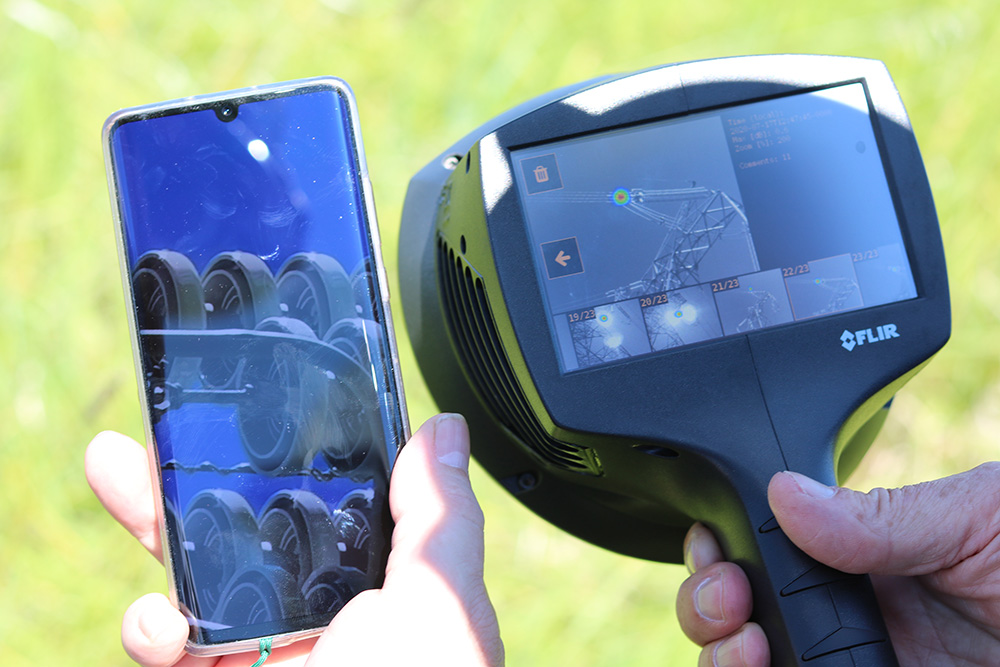 One hand holds the FLIR Si124 Acoustic Camera with an acoustic image of electricity lines on the screen. The other hand holds a smartphone with a close-up digital image of the problem area highlighted on the acoustic image.