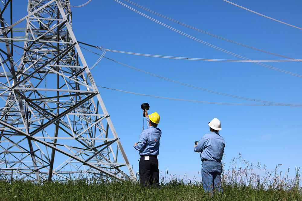 Two men in hardhats stand in front of an electricity pylon. One is pointing the FLIR Si124 Acoustic Imager at the pylon while the other is operating a drone. 