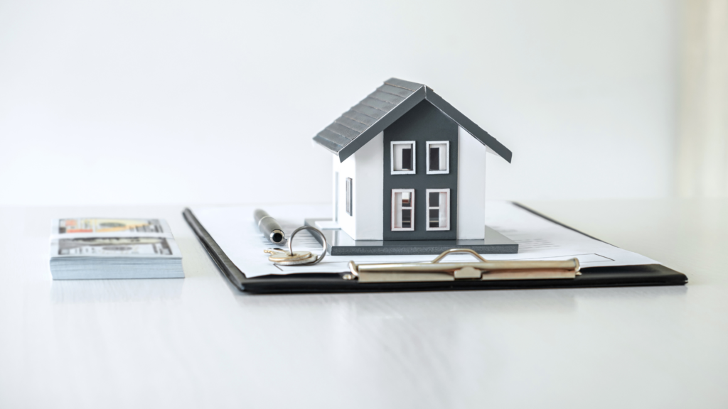 On the left of the image is a neat, small pile of money. Next to it is a clipboard with a contract, keys, pen, and white and grey model house on top of it.