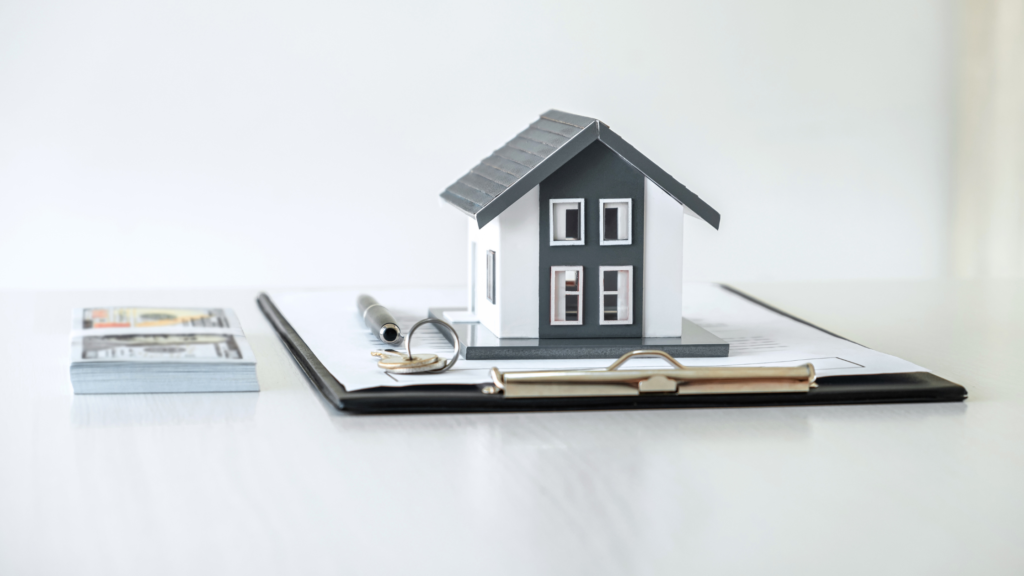 On the left of the image is a neat, small pile of money. Next to it is a clipboard with a contract, keys, pen, and white and grey model house on top of it. 