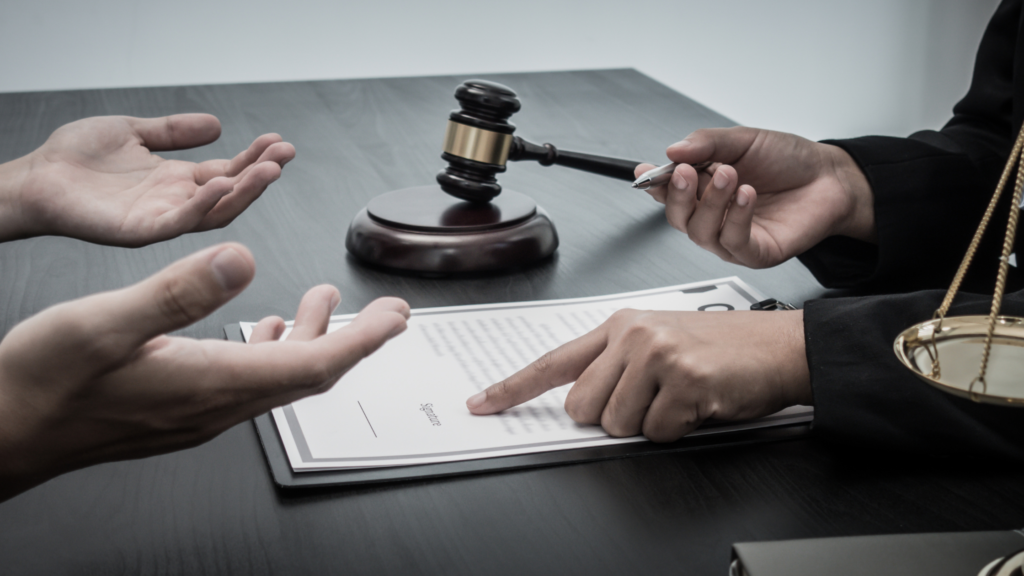 Two people sit across a desk from each other. We can only see their hands. The person's hands on the left of the image are upturned. On the right the person is holding a pen in one hand an using the other to point at a document. Symbols of justice, a judges hammer and scales, are positioned either side of the person on the right. 