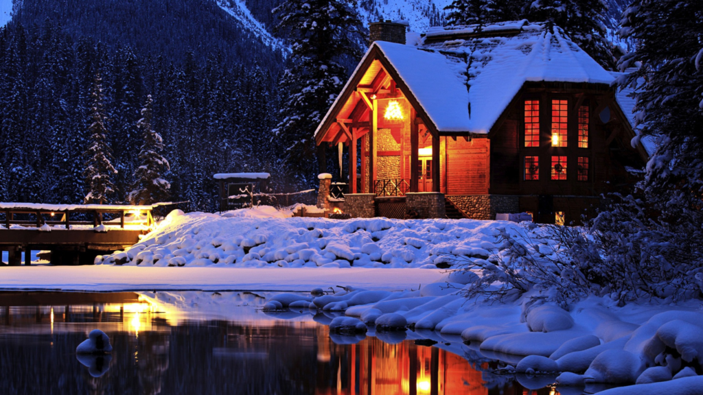 A log cabin illuminated by warm yellow lights stands covered in snow in a Christmas tree forest. It is near a clear lake. 