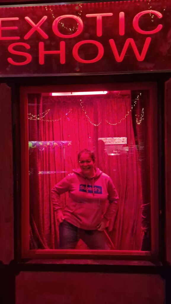 Lynsey strikes a pose in a behind a window illuminated in red. The sign 'Exotic Show' is above it. Lynsey is dressed in jeans and a hoodie. 