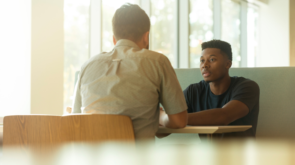 Two men sit in a cafe across the table from each other. The man in a white shirt has his back to the viewer. The man across from him in a black t-shirt is listening intently to what he is saying. 