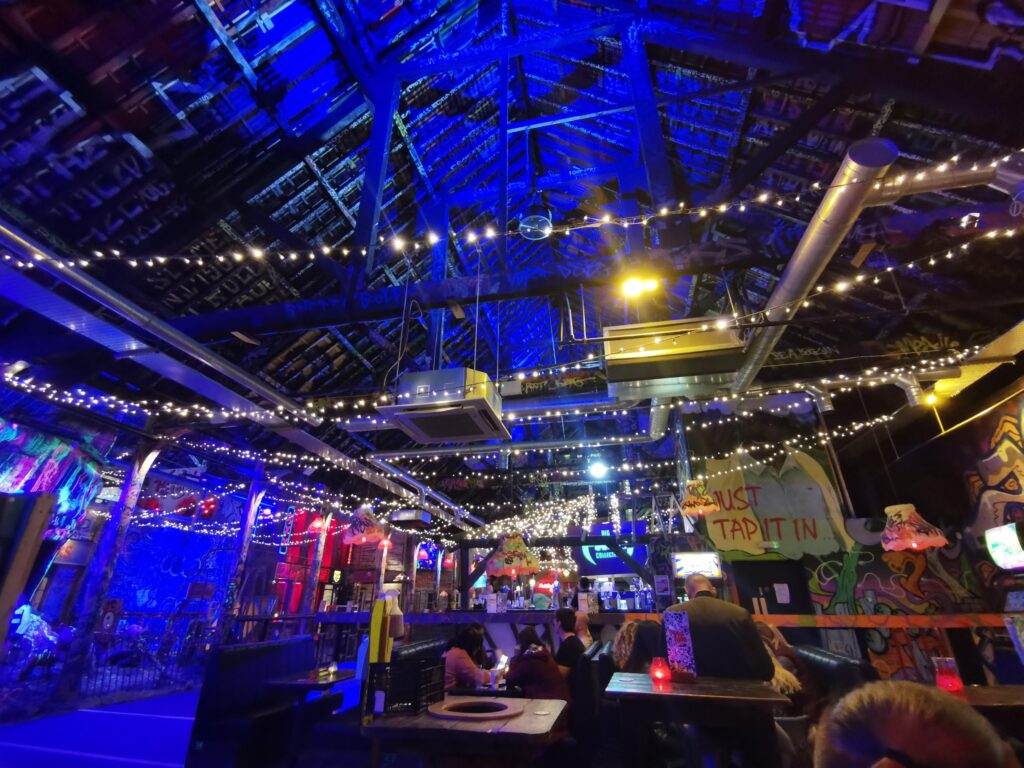 Image of the eating area including tables and benches. The high ceiling is graffitied and the room is illuminated by rows of fairy lights. The room has a look of a warehouse about it. 