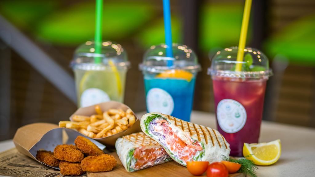 Three smoothies in plastic domed cups stand in the background, on the left is lemon smoothing with a green straw, in the middle a blue one with a blue straw, and on the right is a pink smoothie with a yellow straw. In front of the lemon smoothing fries and breaded nuggets spill out of brown paper fast food packages. In front of the blue smoothie is a smoked salmon wrap, next to it in the right is a slice of lemon, some dill, and three halves of cherry tomatoes. 