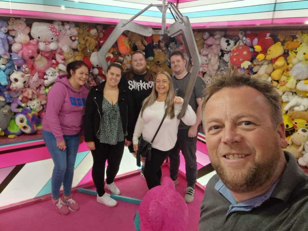 Lynsey, Collette, Adam, Michael, and Clare pose for a photo beneath a grabbing-hook in a life size, pink, pick-up arcade game. The walls around them are filled with teddies. Barry stands at the front. He is taking a selfie with them. 