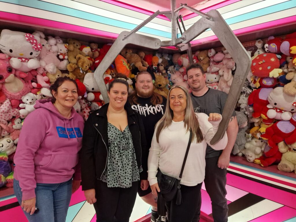 Lynsey, Collette, Adam, Michael, and Clare pose for a photo beneath a grabbing-hook in a life size, pink, pick-up arcade game. The walls around them are filled with teddies. 