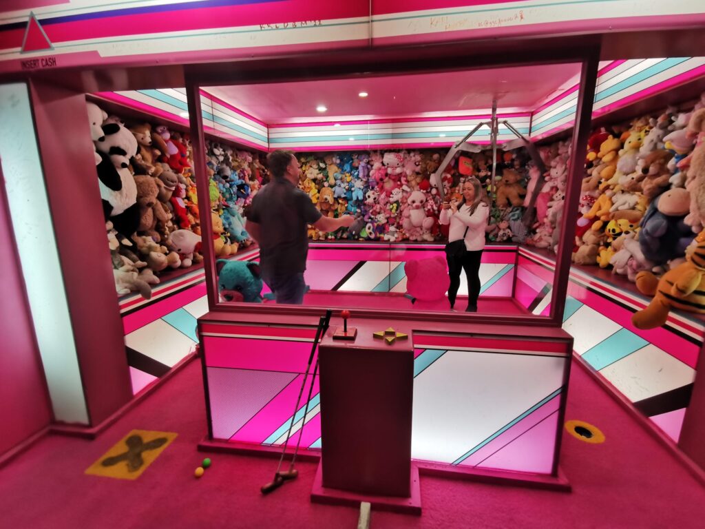 A photo of Clare taking a photo of Barry in a life-size, pink. pick-up arcade game. The walls around them are full of teddies. 