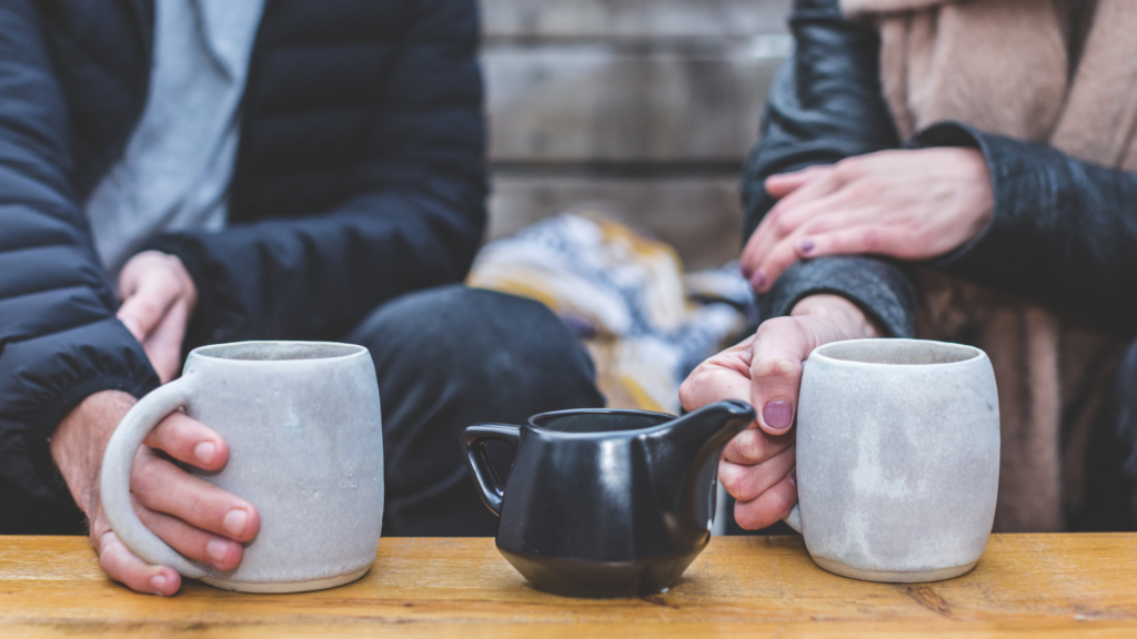 A man and a woman are having coffee outside. Only their hands and torsos are visible. Both hold grey coffee which are standing on a wooden table either side of a black milk jug. 