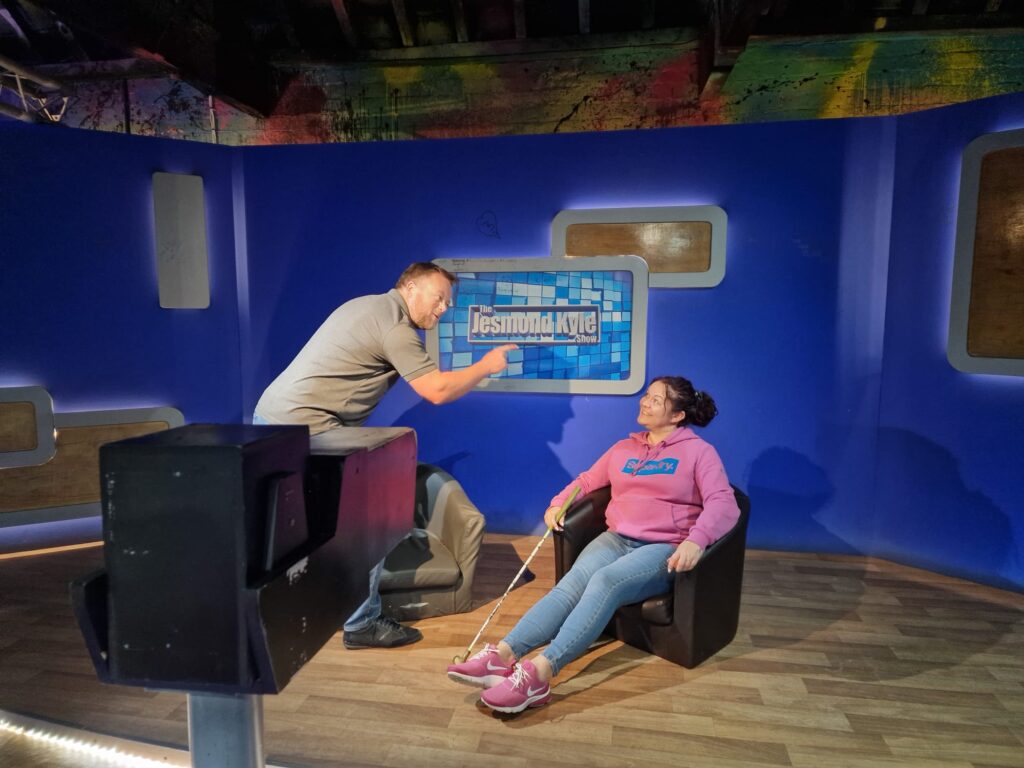 Lynsey and Barry are in a fake Jeremy Kyle set. Barry plays the role of Jeremy Kyle. He leans over Lynsey, who is sitting in an armchair, wagging his finger and pretending to tell her off. 