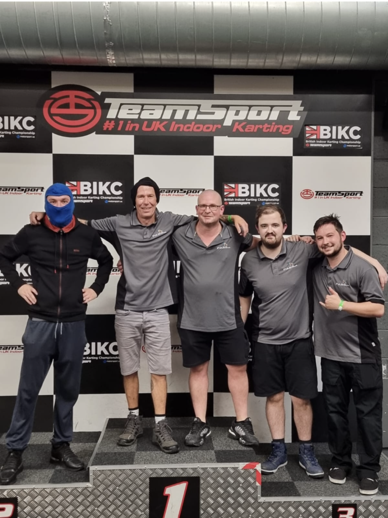 One of the Dispatch karting groups stands on the podium. Four of this team are wearing grey PASS-branded polo shirts and shorts/trousers. The other is dressed in a tracksuit with a blue racing balaclava over his head.  