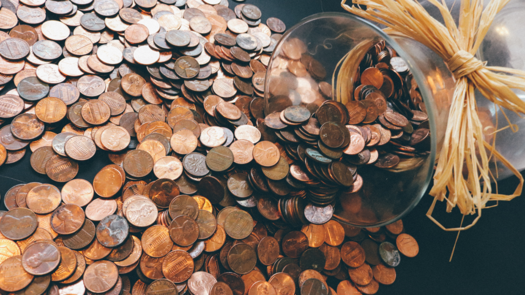 On the right of the image a glass jar with a straw bow has fallen over. Copper coins spill out of it covering the table. 