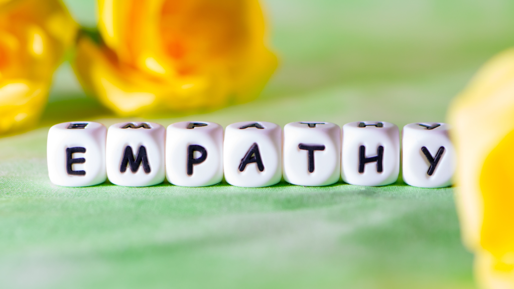White dice with letters on instead of numbers spell out 'Empathy'. They are stood on a table covered in green fabric. Out-of-focus yellow roses are in the background.  