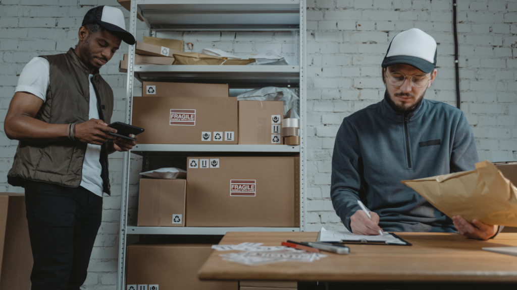 Two men sit are in small office surrounded by boxes and packages. The man on the right sits at a desk. He is holding a brown package and writing something down on a clipboard. The man on the left stands next to a shelving unit filled with boxes. He is checking something on a tablet. 