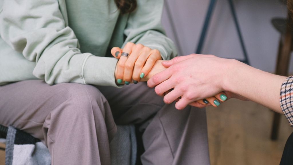 A person on the right holds the hand of the woman on the left between their hands in a comforting gesture. The woman's arms, torso, and legs are visible; only the hands and forearms of the person on the right are visible.