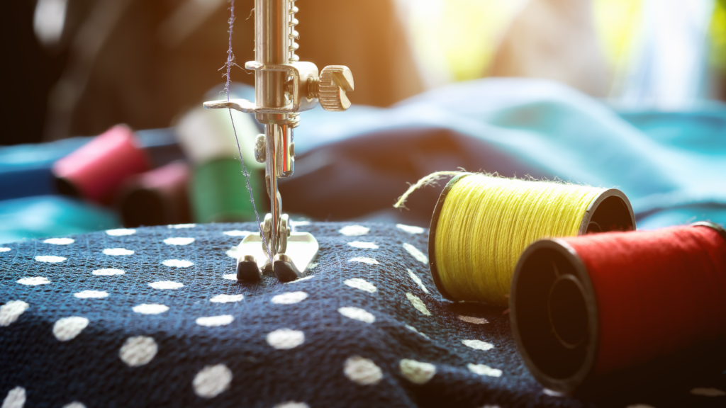 A blue material with white spots is under the foot and needle of a sewing machine. Rolls of red and yellow thread lie next to it. 