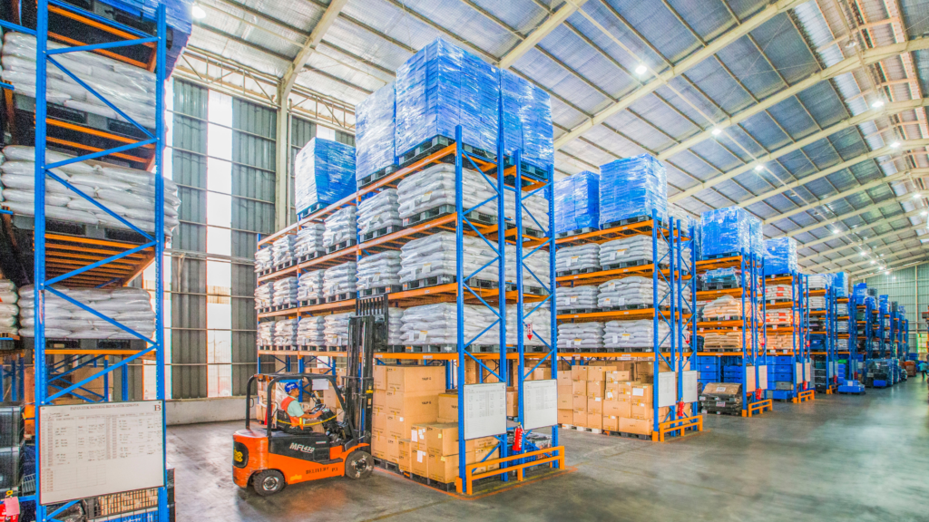 A bright warehouse with a high ceiling and blue and orange shelving units containing boxes and white soil bags. A man in orange high-vis and a blue hardhat is driving an orange forklift truck and removing a pallet of white soil bags from the second shelf. 
