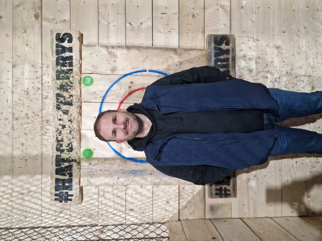 Chris M stands in front of a wooden axe-throwing target. 