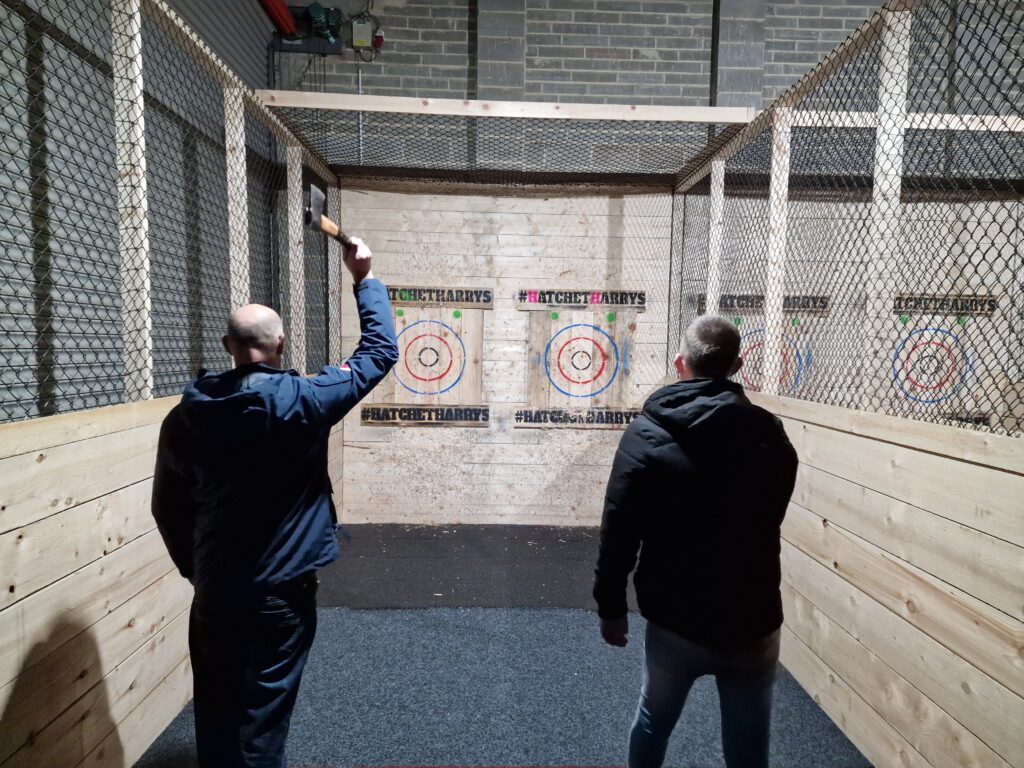 Stephen (left) and Lewis (right) stand with their backs to the camera. A distance in front of them are two wooden axe-throwing targets. Stephen holds an axe in his right hand; it is raised ready to be thrown. 