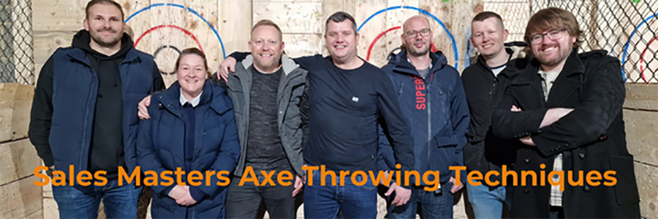 Image of the Sales Team standing in front of two wooden axe-throwing targets. From left to right: Chris M, Sally, David, Gary, Stephen, Lewis, Chris C. At the bottom of the image, large orange text reads "Sales Masters Axe Throwing Techniques". 