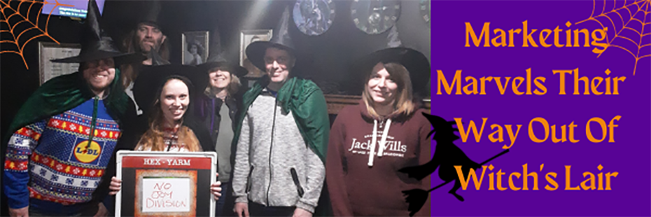 At the left of the image is a picture of the marketing team posing in the Hex escape room. From left to right: Calum, Stacey, Jess, Katie, Stuart, Rachael. Jess is holding a board with the team's name, "No Joy Division", in the centre above Cluedini's name and logo. On the right of the image, large orange text on a purple background reads "Marketing Marvels Their Way Out Of Witch's Lair". In the top left and right corners of the image are orange spiderwebs. At the bottom left of the orange text is a black, cartoon witch flying on a broomstick. 