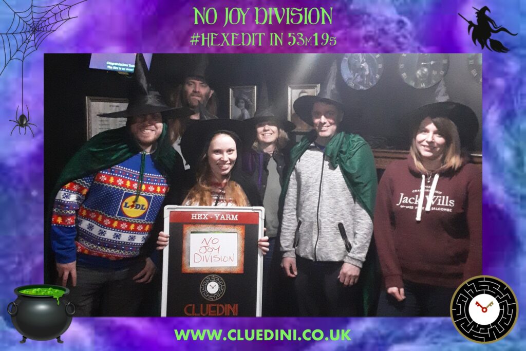 The marketing department pose for a photograph in the Hex escape room. From left to right: Calum, Stacey, Jess, Katie, Stuart, Rachael. Jess is holding a board with the team's name, "No Joy Division", in the centre above Cluedini's name and logo. The photo is framed by a swirling blue and purple background. At the top and in the centre of the background, green text reads "No Joy Division #HEXEDIT In 53m 19s". At the bottom and in the centre of the background, green text reads "WWW.CLUEDINI.CO.UK". In each corner is a cartoon image. In the top left there is a spider handing from a web; in the top right is a witch flying on a broomstick; in the bottom left is a cauldron with a green potion bubbling inside it; in the bottom right is the Cluedini logo.