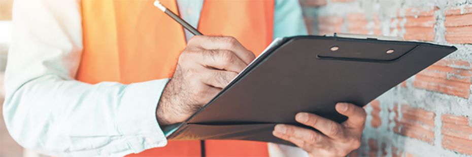 The torso of a man in a white shirt and orange high-vis gilet is visible. He stands next to a red brick wall and is using a pencil make notes on a black clipboard. 