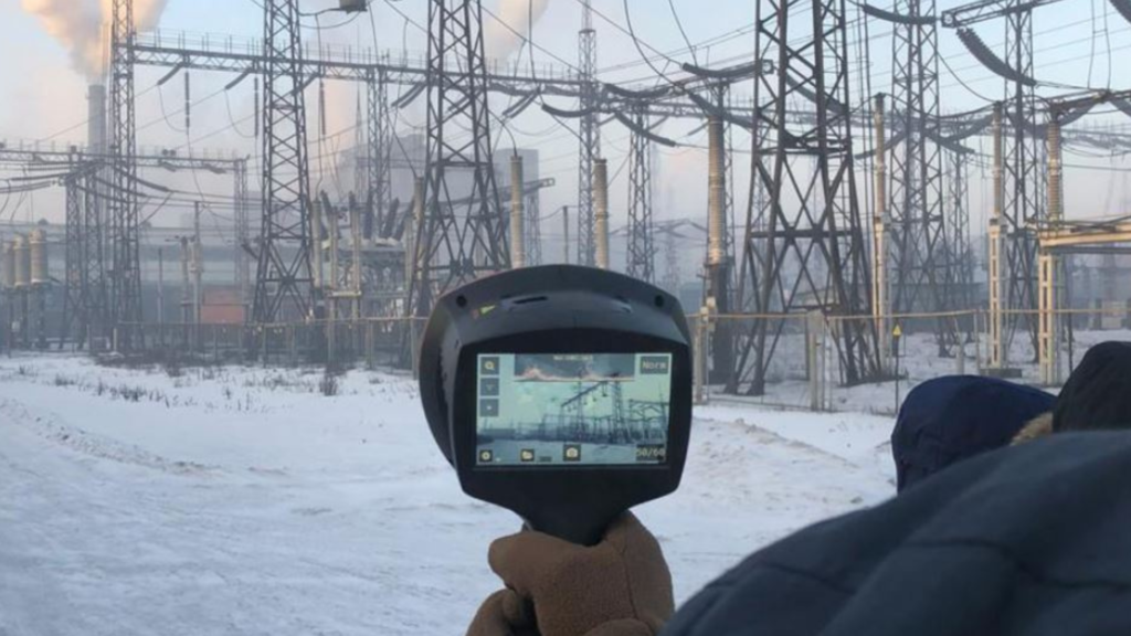 A man holding the Teledyne FLIR Si124 Acoustic imager pointing it at a collection of electricity pylons. The plant is covered in snow.  