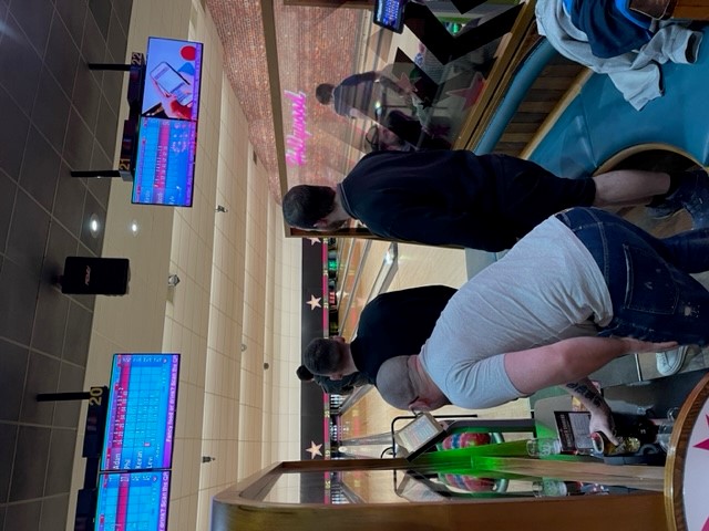 The Dispatch team stand looking down a bowling alley. Phil bends to pick up his drink. 