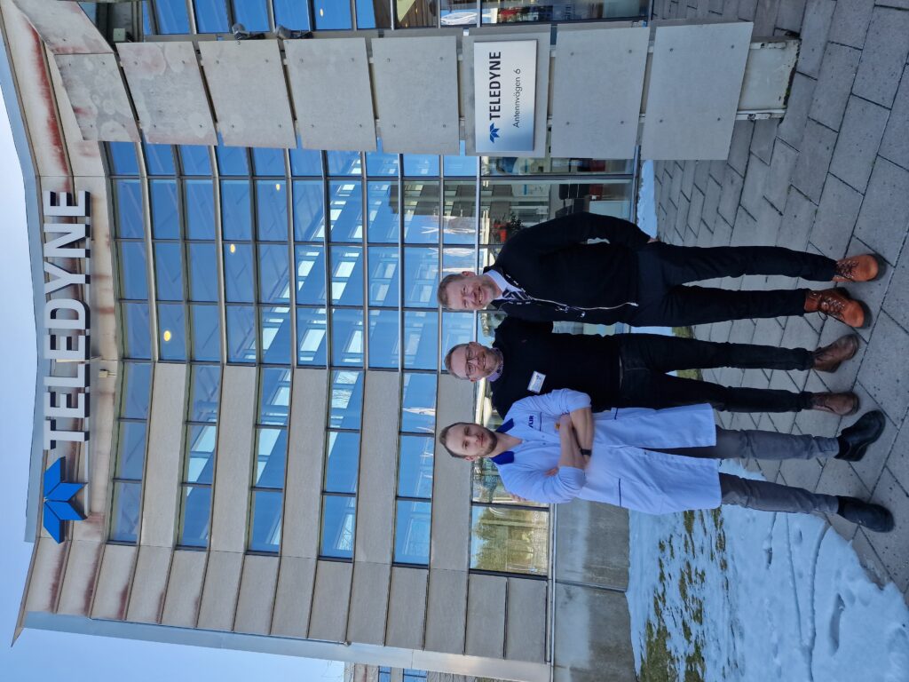 From left to right Thomas Hansson, David Atkins, and Jonas Bolinder stand on a snowy path outside a tall glass and stone building with a big Teledyne sign at the top.  