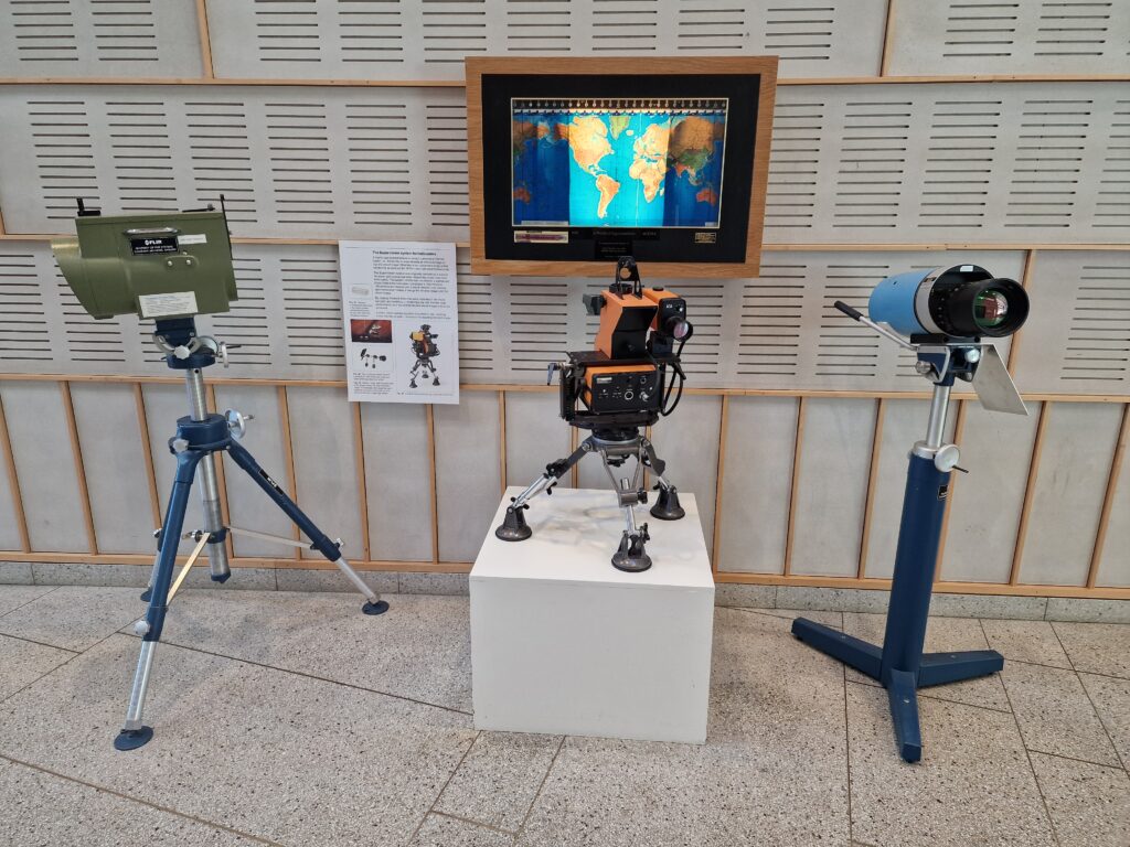 Three early Teledyne camera models. The ones on the left and the right are mounted on tripods. The one in the centre is mounted on a small tripod which, in turn, is stood on a white platform. In the background is a US map of the world. 