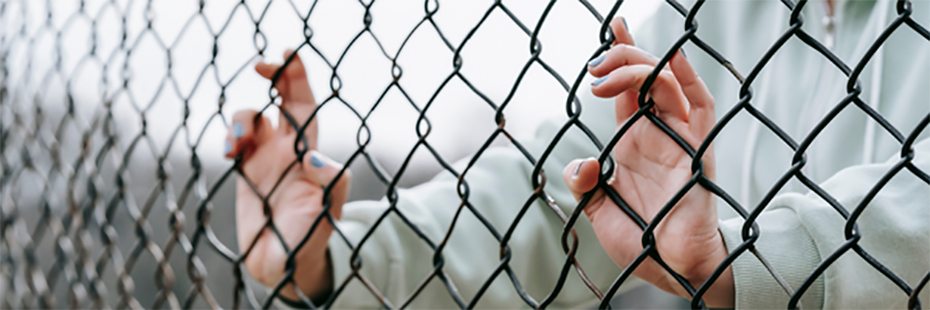 A woman holds onto a mesh fence. Only her hands, arms, and torso can be seen. 